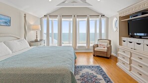A bedroom with a large bed, a ceiling fan, a TV, and a view of the ocean through large windows.