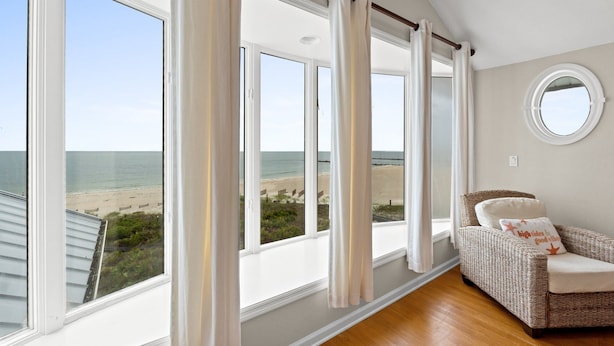 A room with large windows overlooking a beach, a wicker chair with a cushion, and a round mirror on the wall.
