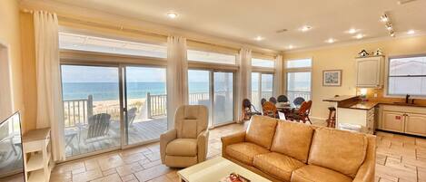 A spacious living area with a large window overlooking the beach, a dining table with chairs, a sofa, and a kitchen area.