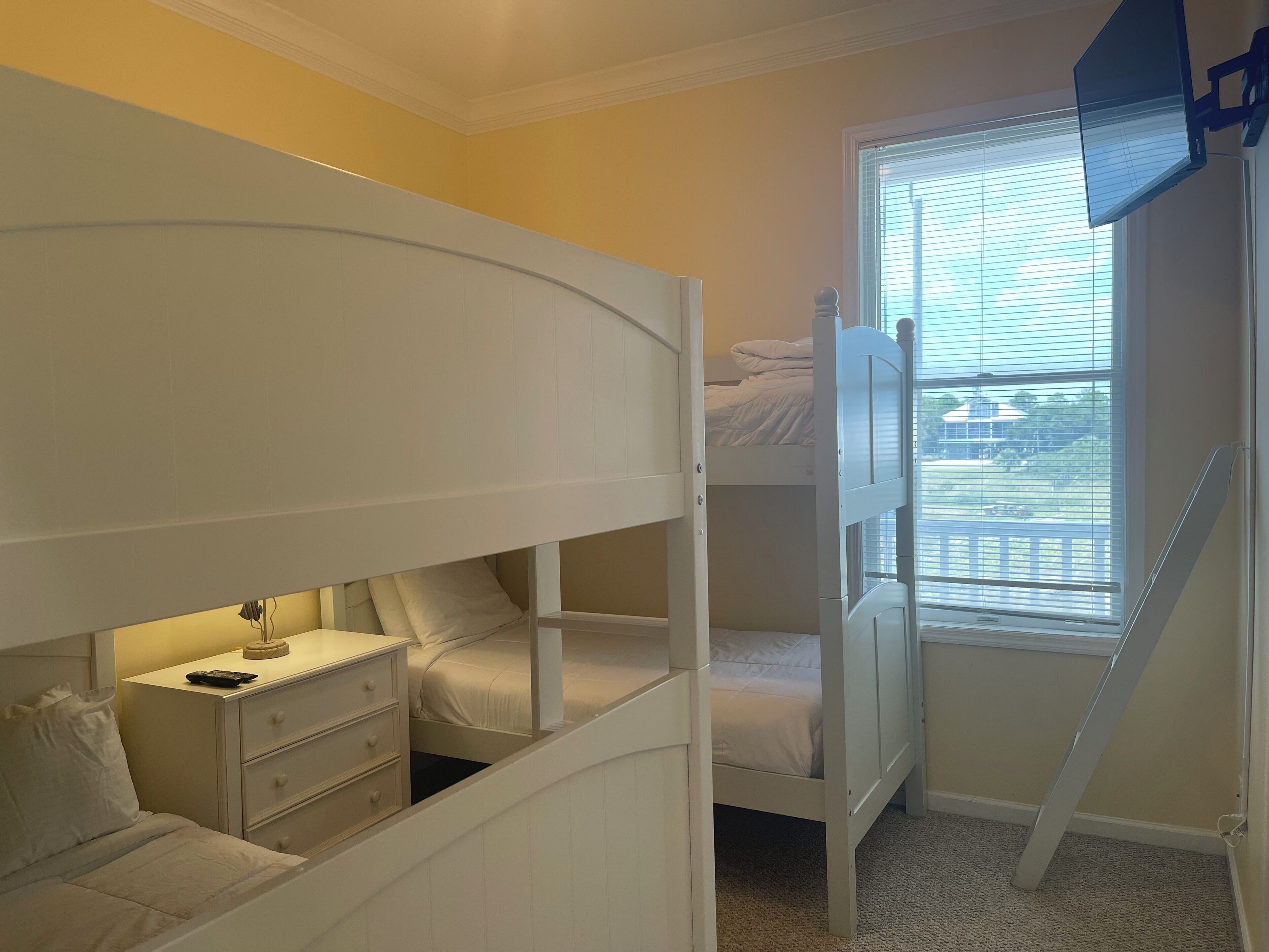 Une chambre avec des lits superposés, une table de chevet avec une lampe et une fenêtre avec des stores.