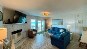 A living room with a blue sofa, a fireplace, a television, and a view of the beach. - Ocean Mile F-4, St. George Island, Community Pool, 2 BR (St. George Island)