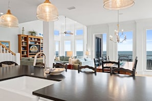 A spacious living area with a large kitchen island, dining table, and chairs. There is a bookshelf with decorative items and a view of the ocean through large windows. - Southern Splendor, BF, Private Pool, 5BR (St. George Island)