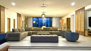 Un saló modern amb un sofà modular, un reposapius i una cadira blava i singular. Hi ha una zona de dinar amb taula i cadires, i una cuina amb armaris de fusta i electrodomèstics.