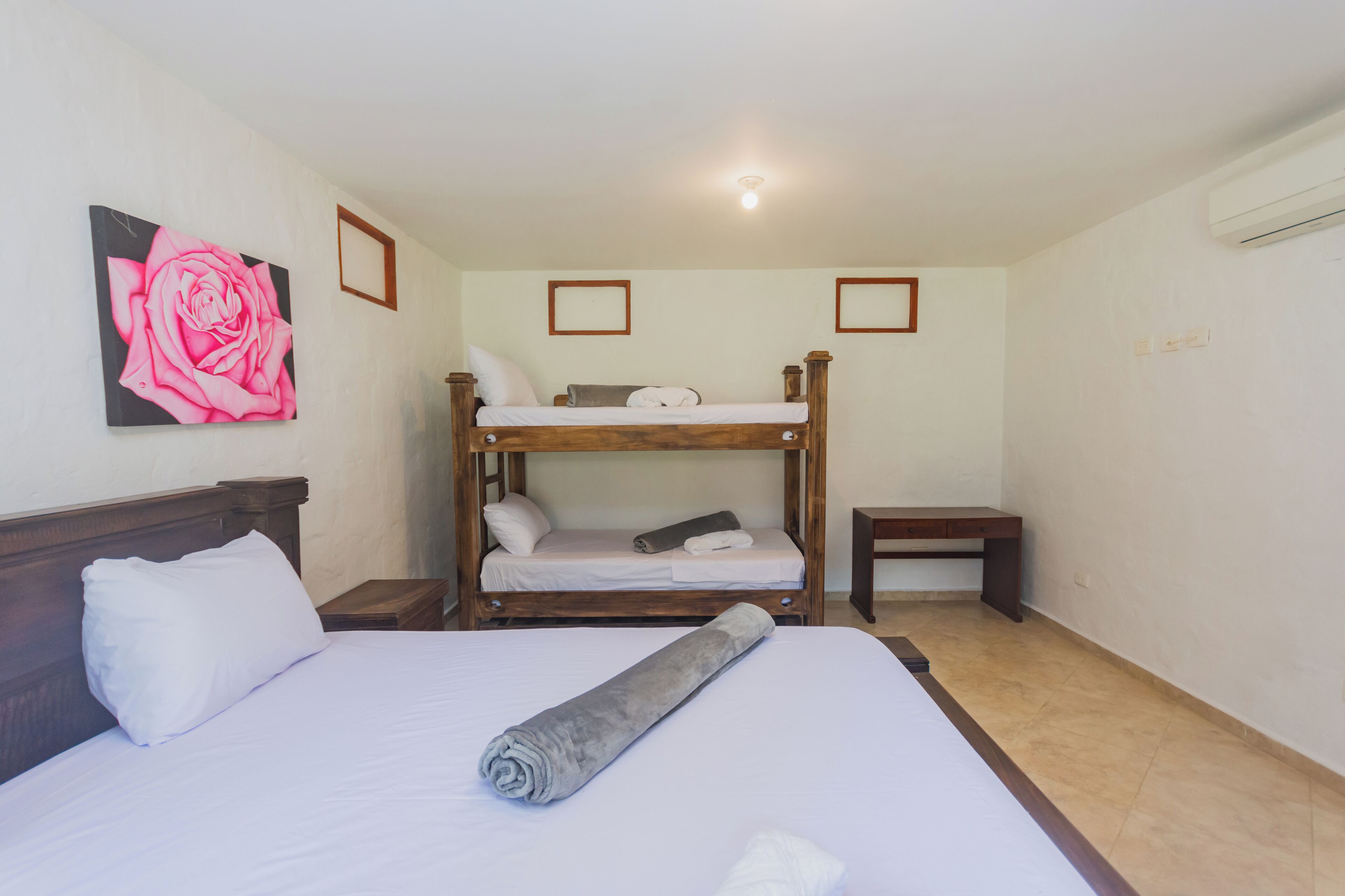 A room with a bunk bed, a single bed, a rolled-up towel, and a painting of a rose on the wall.