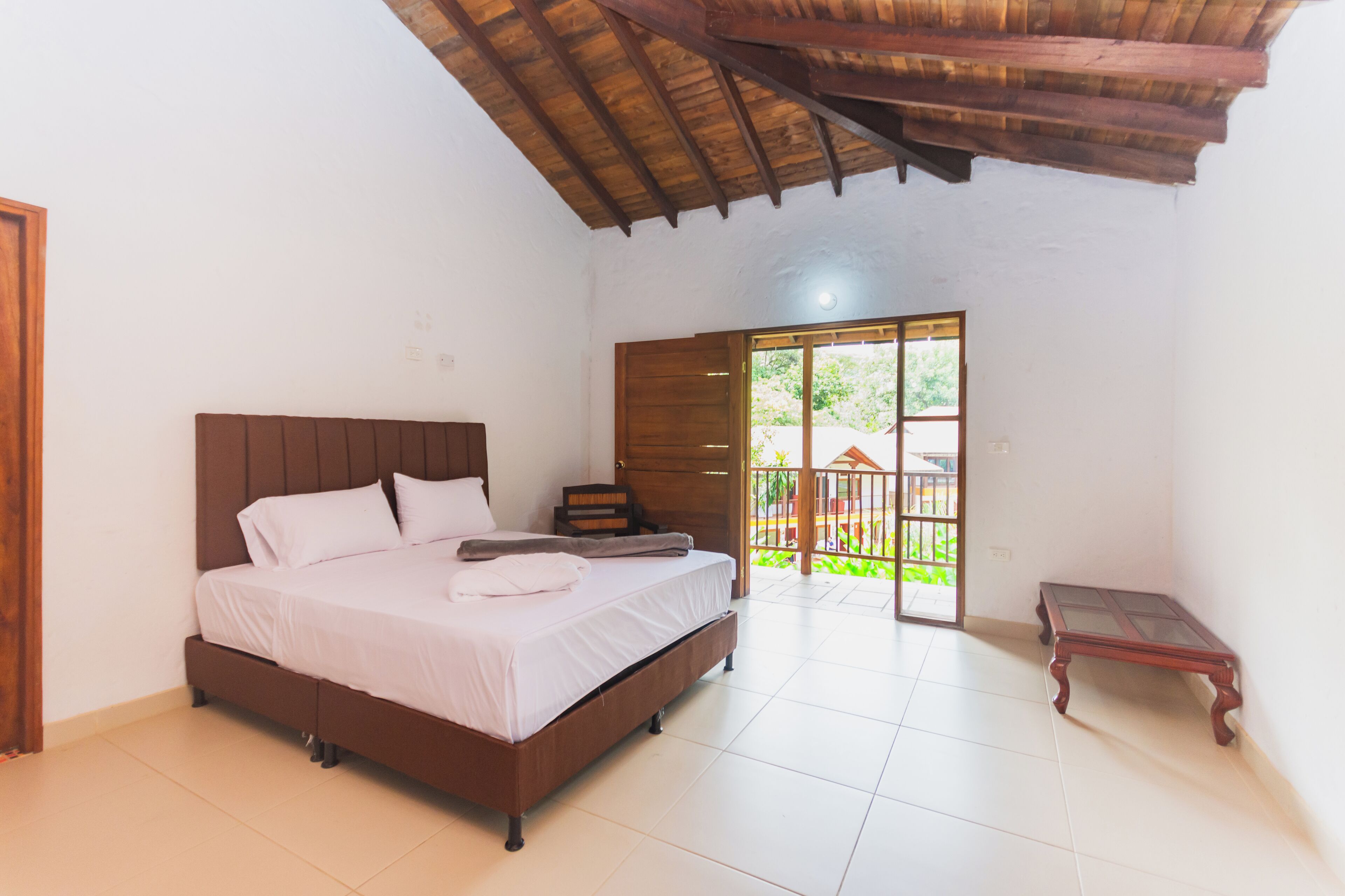 A bedroom with a wooden bed, a small table, and a view of the outside through a door.