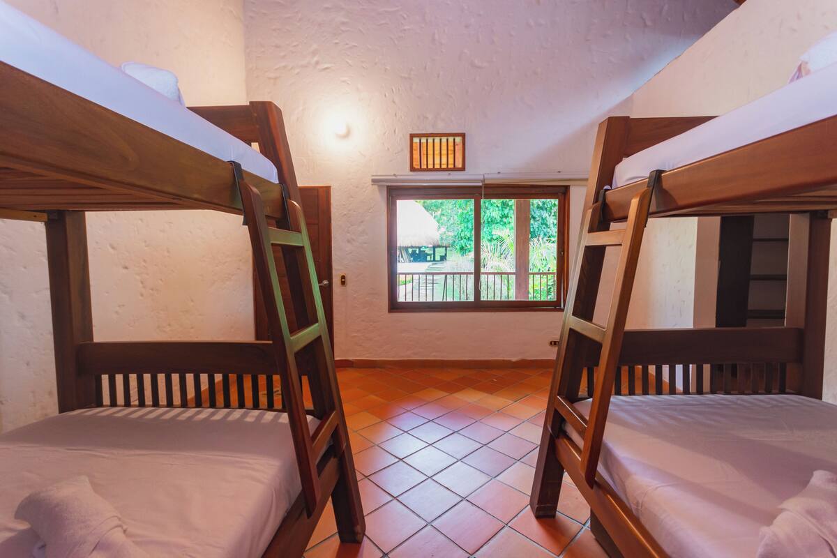 A room with two bunk beds, a window with a view of greenery, and a small light fixture on the wall.