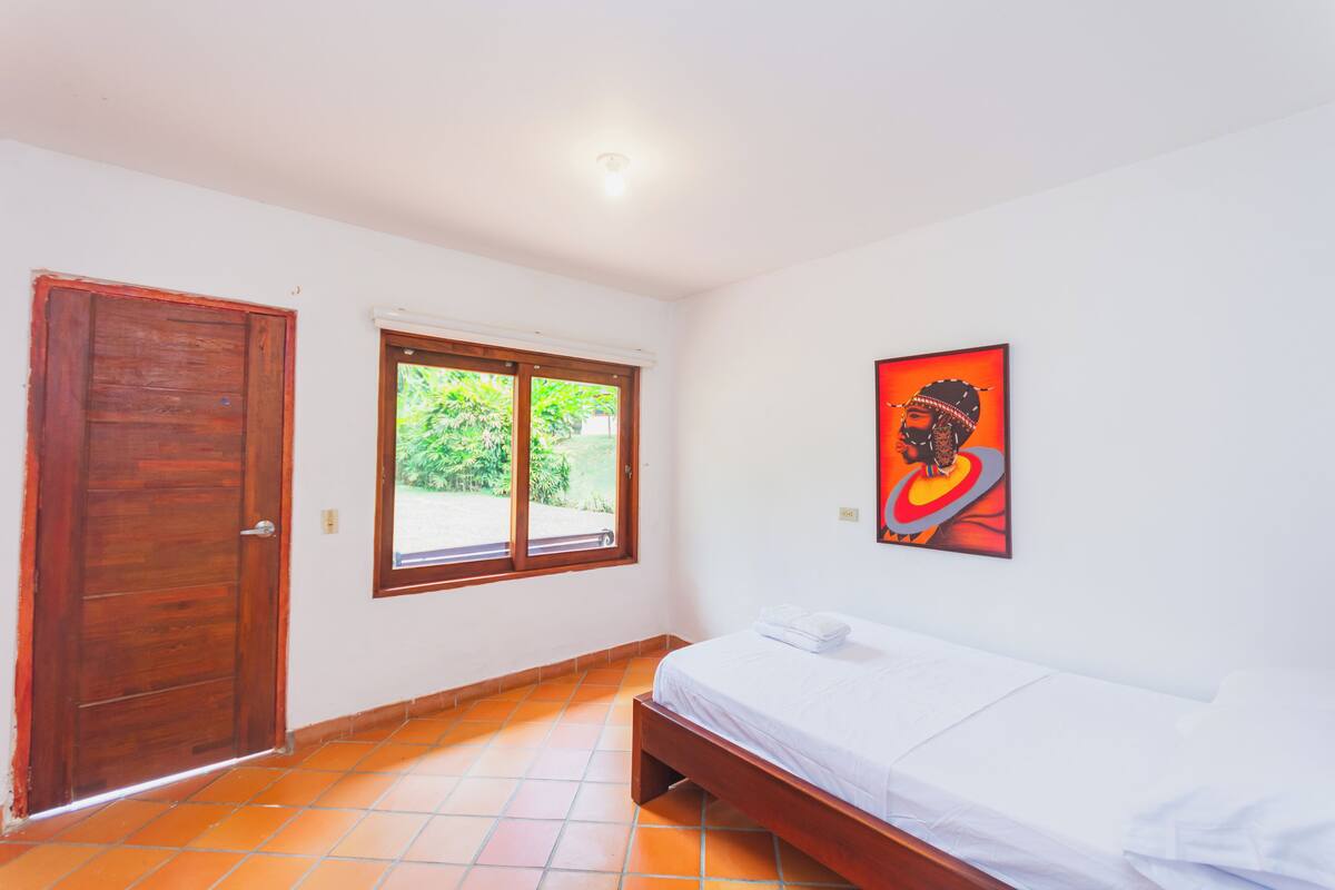 A bedroom with a wooden door, a window with a view of greenery, and a bed with white bedding.
