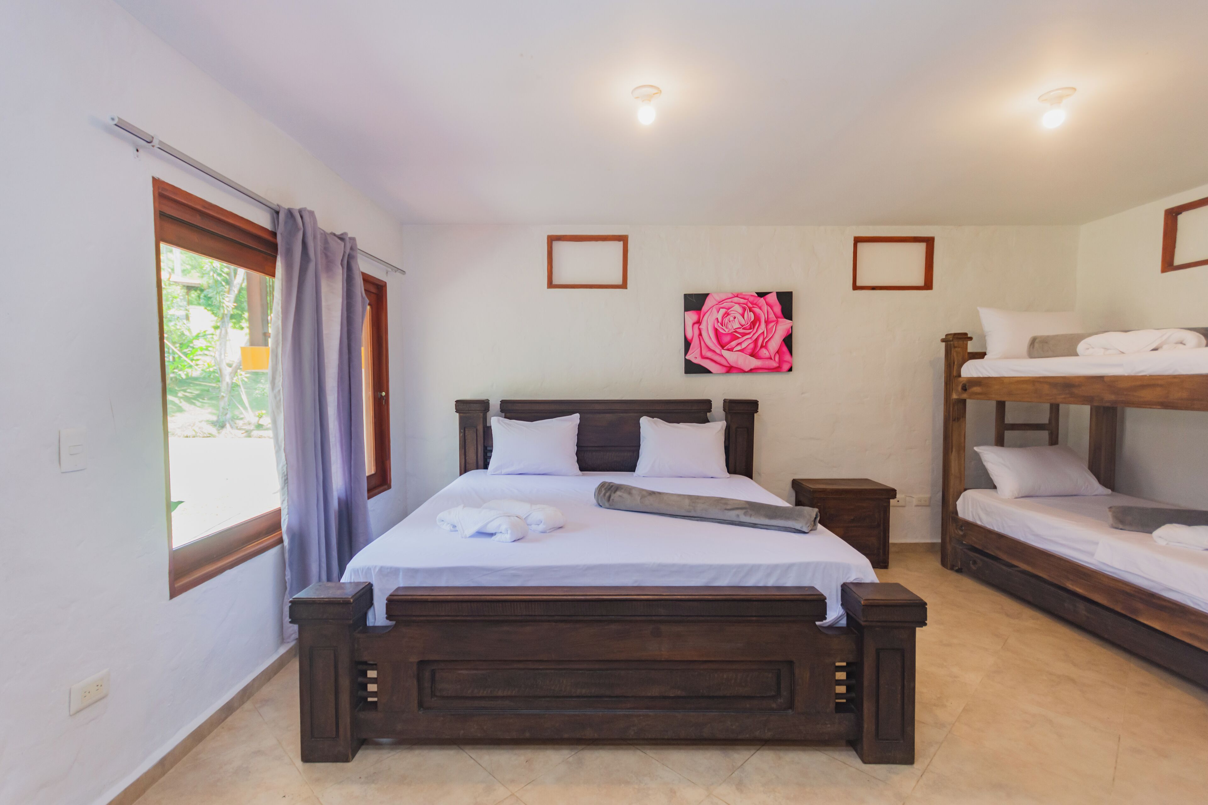 A bedroom with a large bed, a smaller bunk bed, a wooden headboard, a nightstand, and a window with curtains.