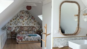 Et soveværelse med skrå vægge, en seng med blomstret sengetøj, en træstige, et hvidt skab, et stort spejl og et badeværelse med en hvid vask og flisebeklædt væg.