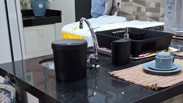 A kitchen with a black countertop, a sink, a stove, and a microwave. There is a blue bucket on the countertop and a striped cloth on the floor. - Ap Próximo ao IME UFRJ e Pão de Açúcar (Rio de Janeiro)