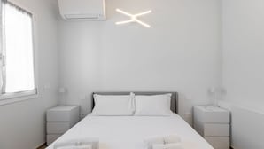 Et minimalistisk hotelværelse med en stor seng, hvide sengetøj, to sengeborde med lamper og en klimaanlæg på væggen.