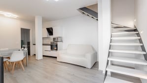 En moderne stue med en hvid sofa, et spiseområde med bord og stole, et køkken med hårde hvidevarer, og en trappe.