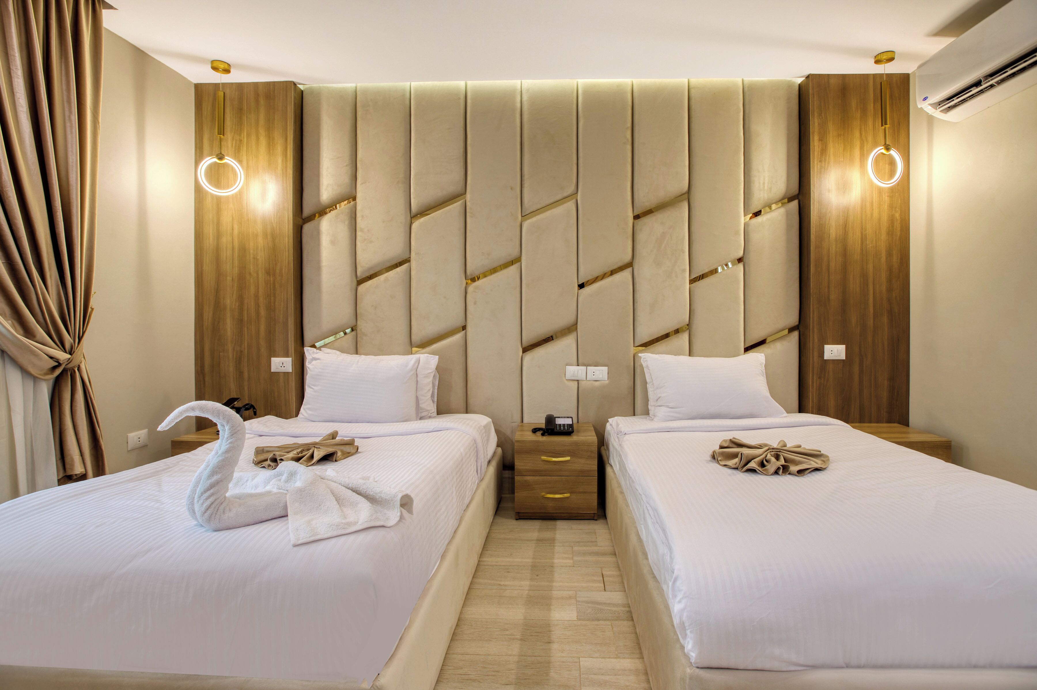 Ein Hotelzimmer mit zwei Betten, einem hölzernen Kopfteil, Nachttischen mit Lampen und einem Vorhang auf der linken Seite.