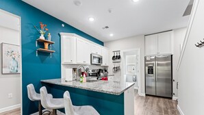 A modern kitchen with white cabinets, a teal accent wall, a central island with bar stools, stainless steel appliances, and a hanging pot rack. - Luxury Condo / Pool / Game Room / Grill / Pets (Branson)