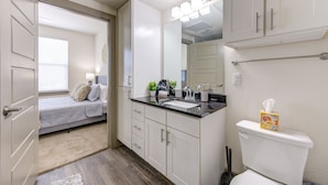 A bathroom with a toilet, sink, and mirror. There is a bed with a window and a door leading to another room.