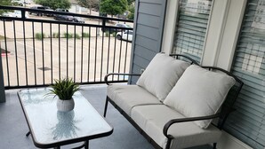 A balcony with a sofa and a table, a potted plant, and a view of a parking lot.