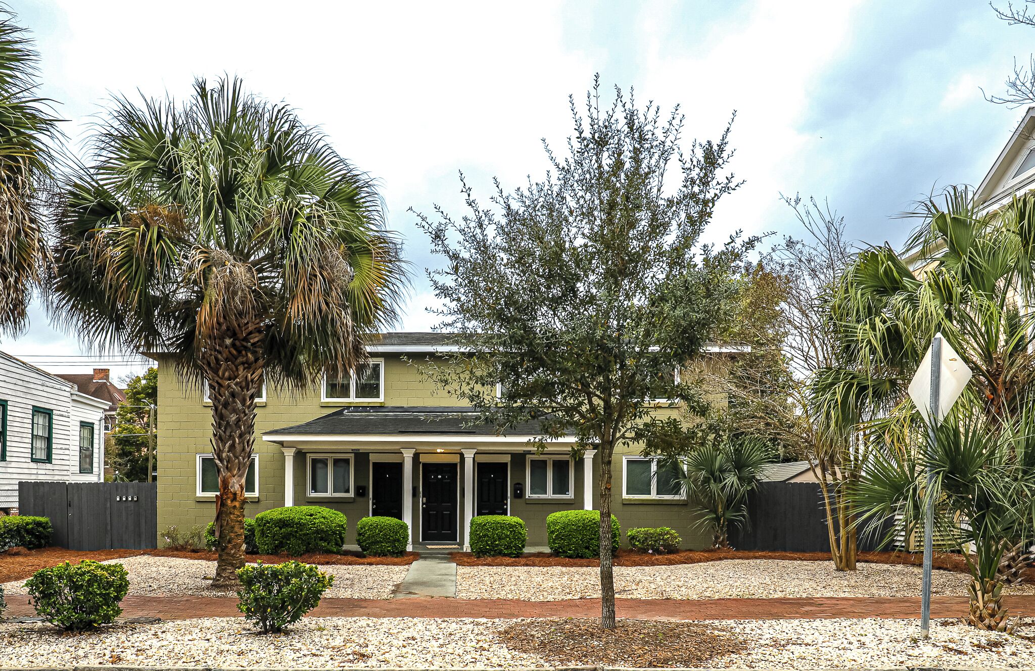 A two-story house with a front porch, surrounded by a gravel driveway and various shrubs.