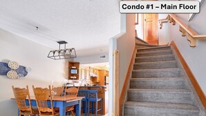 Un coin repas avec une table en bois et des chaises bleues, une cuisine avec des meubles en bois et un escalier recouvert de moquette.