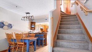 A dining area with a wooden table and blue chairs, a kitchen with wooden cabinets, and a carpeted staircase.