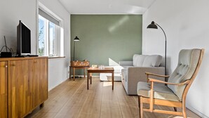 A compact living space with a wooden cabinet, a television, a green accent wall, a grey sofa, a wooden armchair, and a small table with a lamp. - Awesome home in Tønder with WiFi (Tønder)