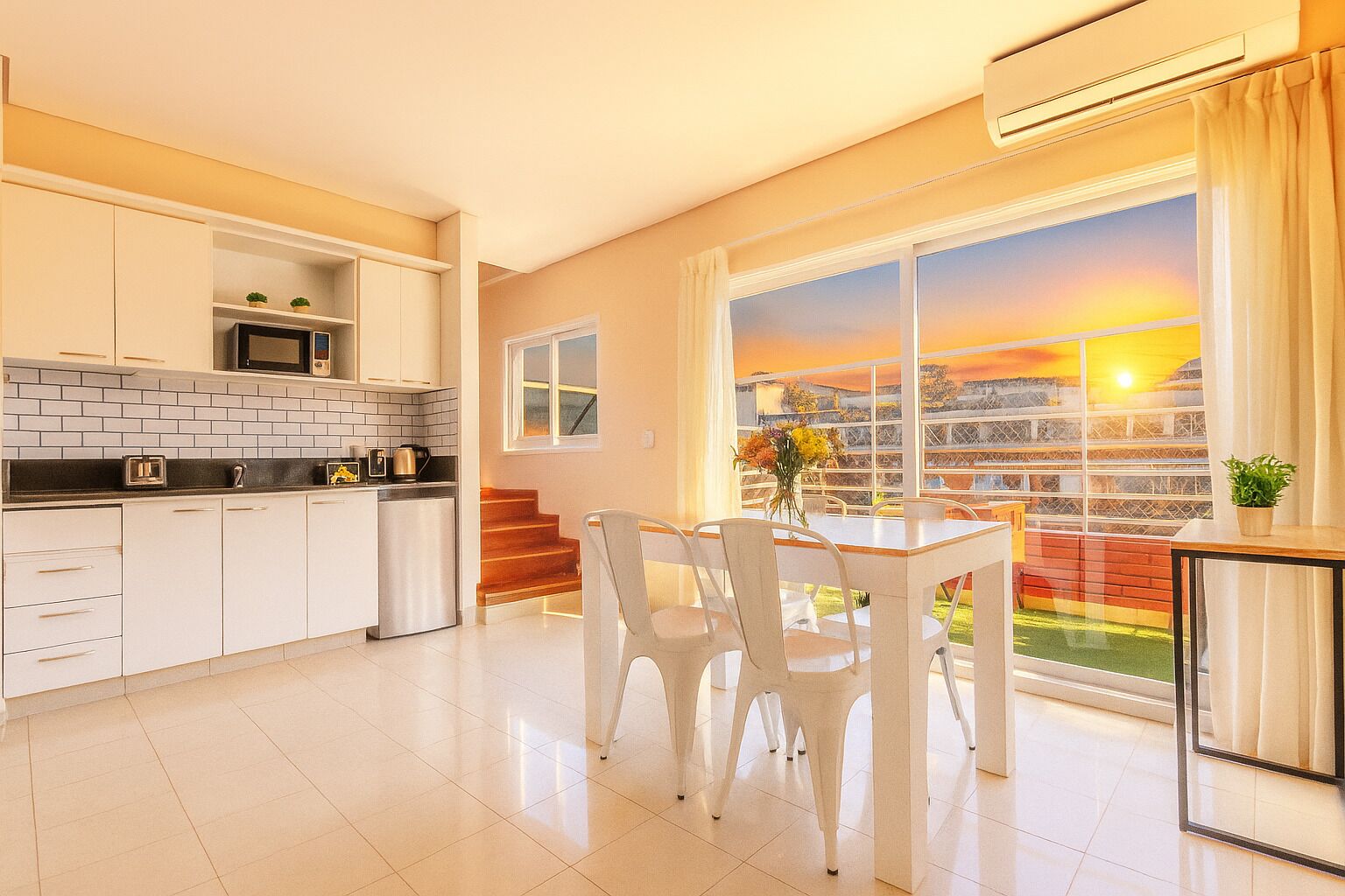 Cocina moderna con armarios blancos, encimera negra y refrigerador de acero inoxidable. Hay una zona de comedor con sillas blancas y una mesa, y un ventanal con vistas a un atardecer.