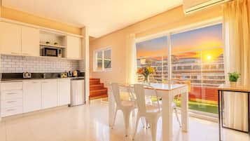Cocina moderna con armarios blancos, encimera negra y refrigerador de acero inoxidable. Hay una zona de comedor con sillas blancas y una mesa, y un ventanal con vistas a un atardecer.