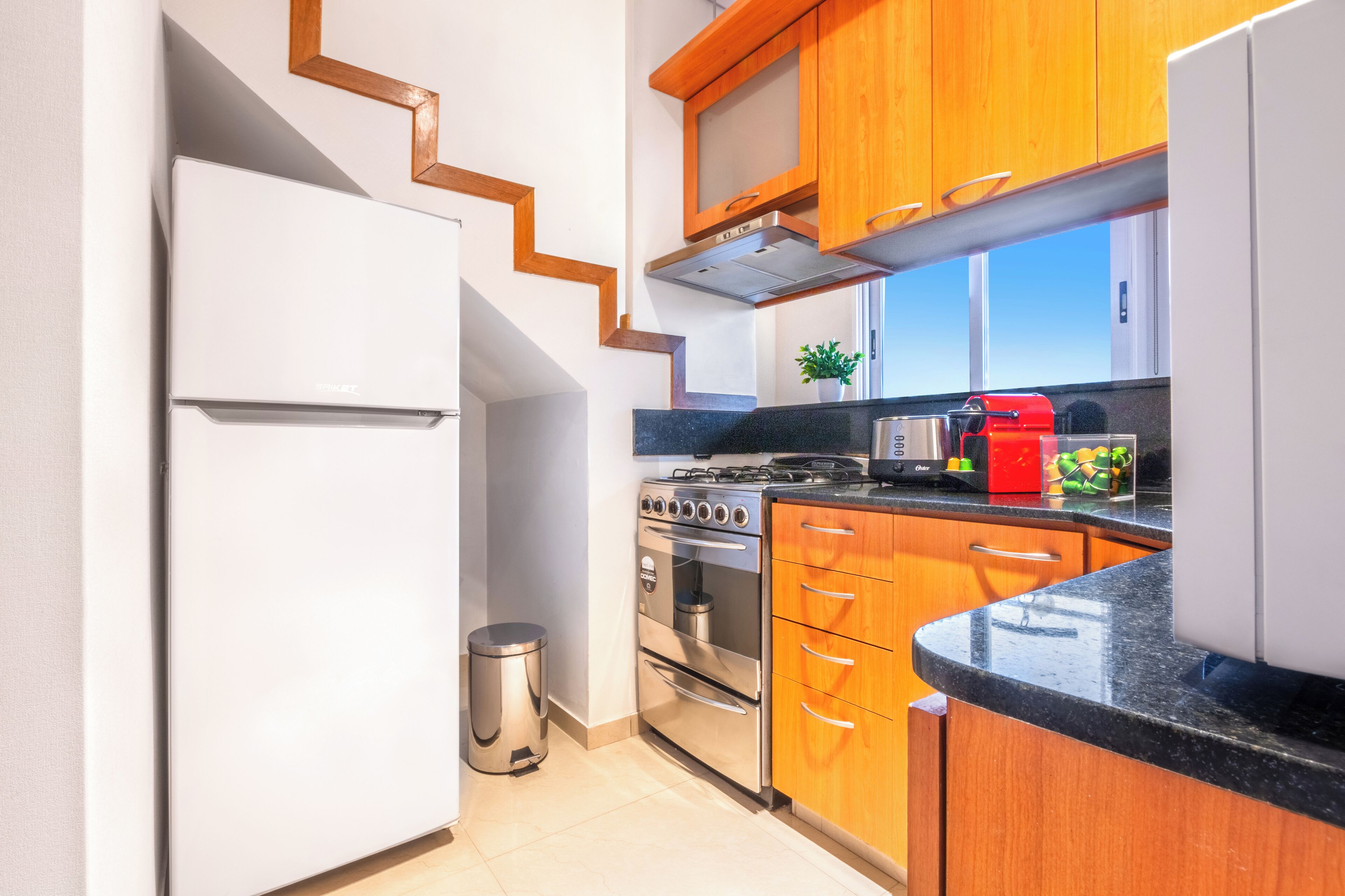 A kitchen with wooden cabinets, a stainless steel refrigerator, a stove, and a trash can.