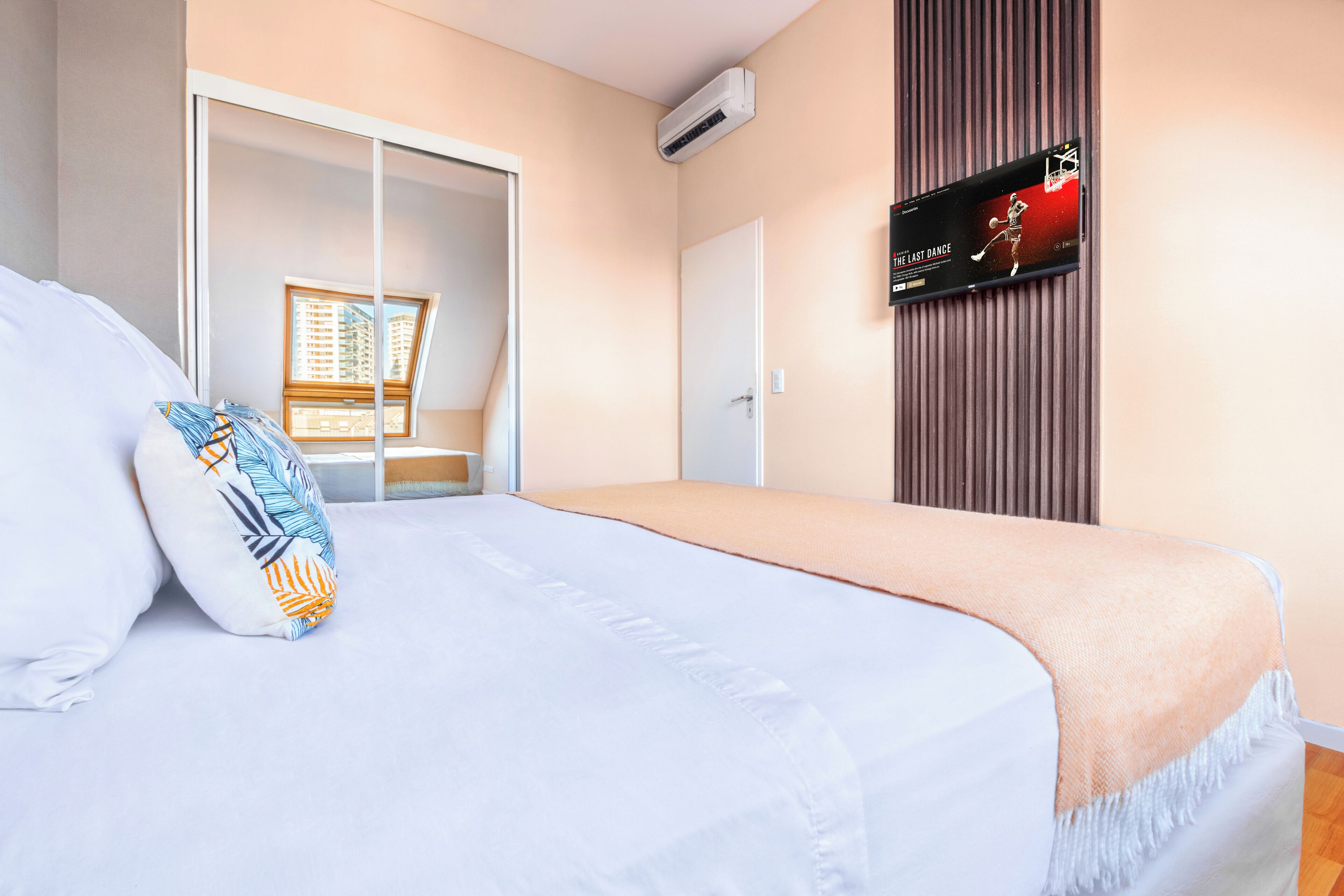 A hotel room with a bed, pillows, a TV on the wall, and a sliding door.