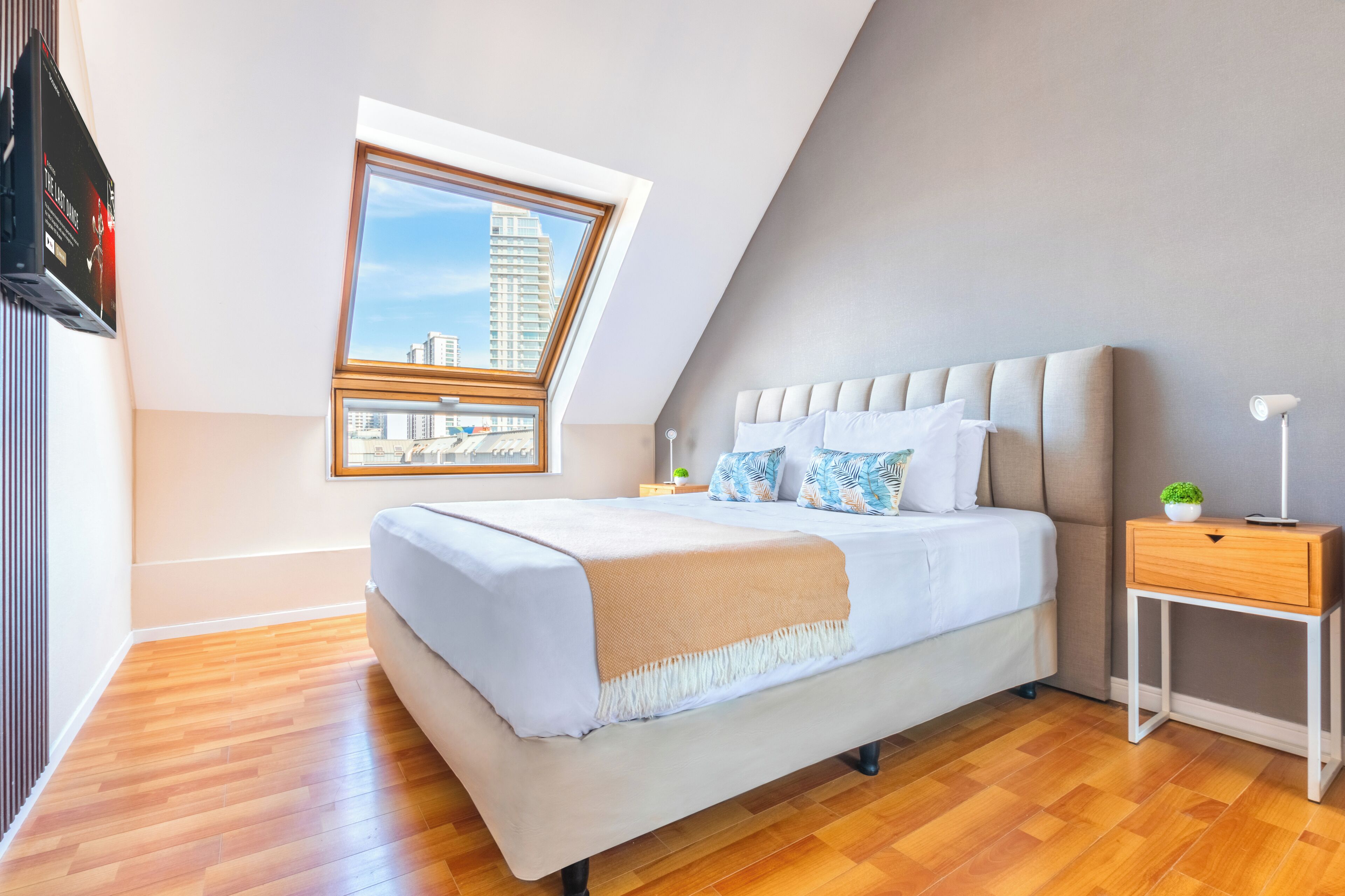 Un dormitorio moderno con techo inclinado, una cama grande con manta color beige, una mesita de noche con lámpara y una ventana con vista a la ciudad.