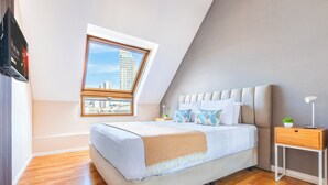 A modern bedroom with a sloped ceiling, a large bed with a beige blanket, a nightstand with a lamp, and a window with a city view. - Modern Lofts in Puerto Madero (Buenos Aires)