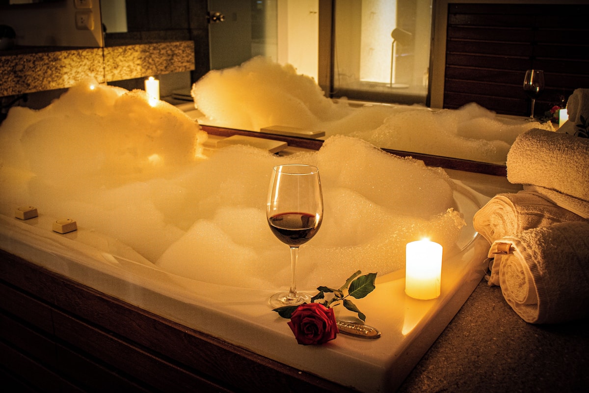 A bathtub filled with bubbles, a glass of red wine, a lit candle, and a rolled towel.