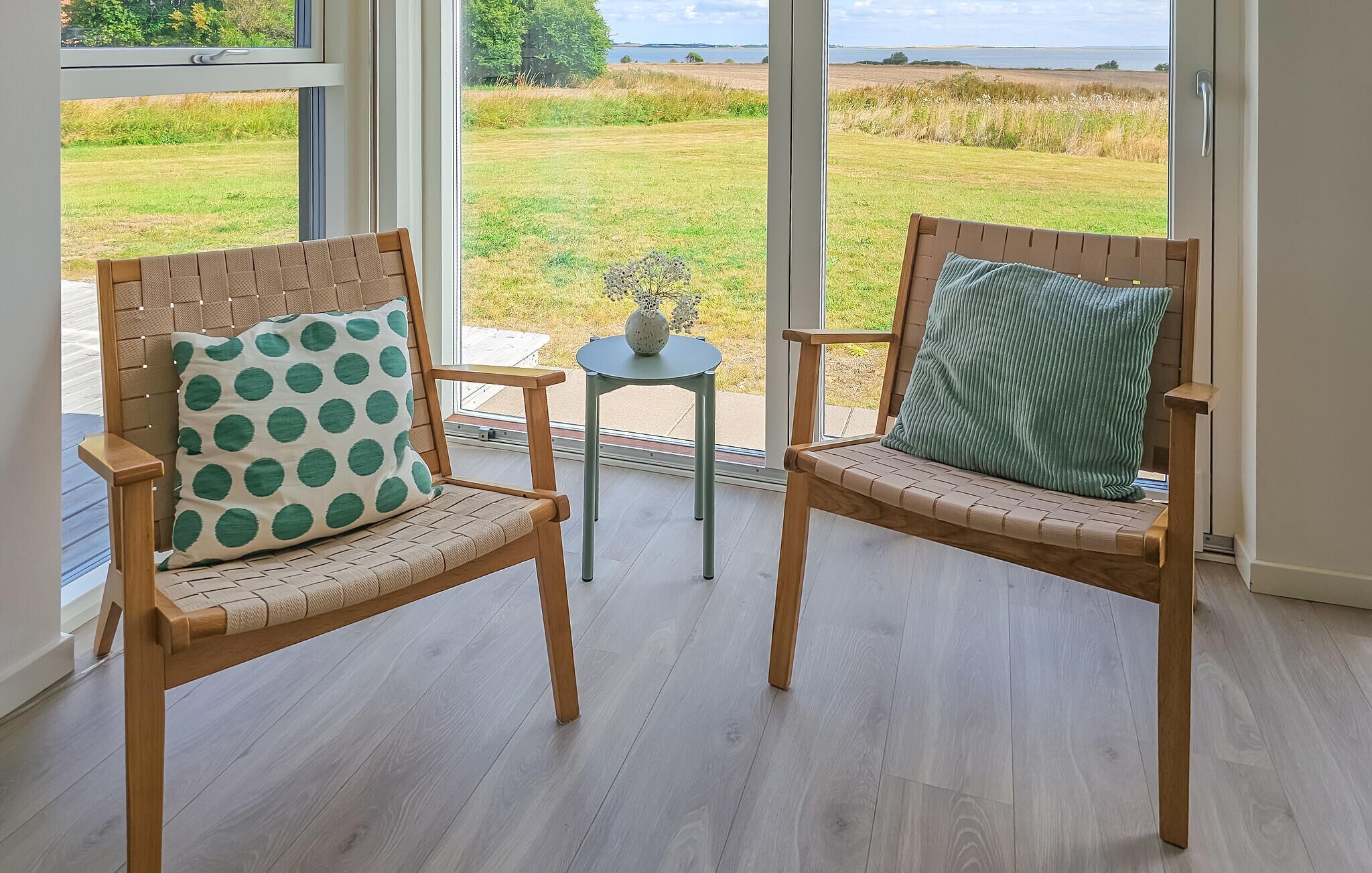 Two wooden chairs with cushions, a small table, and a view of a grassy field through large windows.