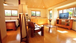 A spacious living area with a wooden floor, a sofa, a coffee table, a TV, and a kitchen with wooden cabinets and a window.