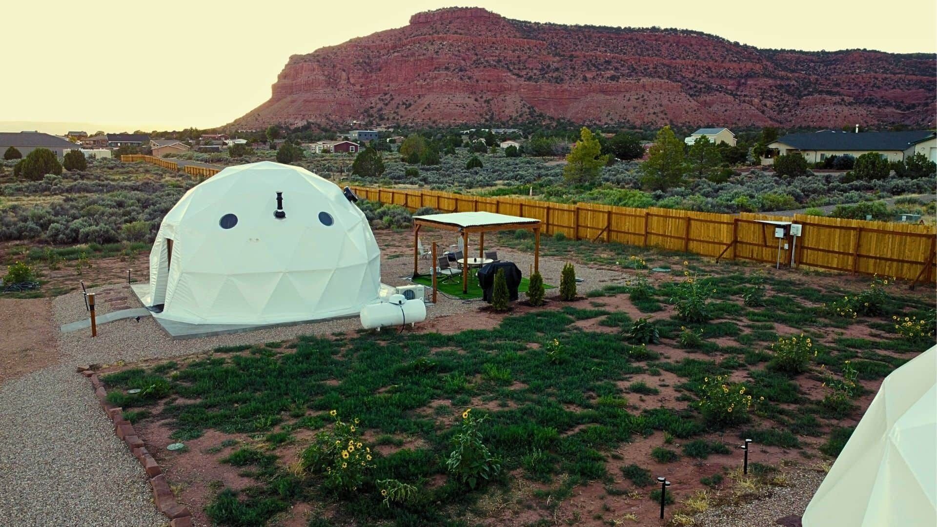 Une structure en forme de dôme géodésique dotée d’un patio, entourée d’une cour clôturée avec une végétation clairsemée et, en arrière-plan, d’une montagne.