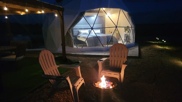 Eine geodätische Kuppel mit einem Bett, zwei Adirondack-Stühlen und einem Lagerfeuer.