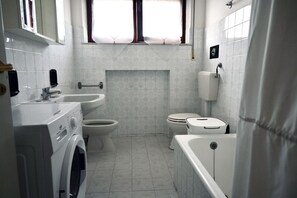 A compact bathroom with a washing machine, two toilets, a bathtub, and a window with curtains.