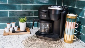 Keurig-kahvinkeitin keittiön työtasolla, sen vieressä kahvikuppialusta, ruukkukasvi ja koristeellinen muki.