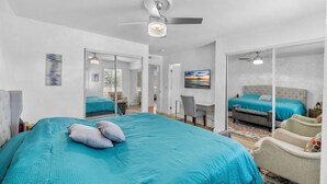 Um quarto espaçoso com uma cama grande, uma escrivaninha e uma cadeira. Há um ventilador de teto e uma TV fixada na parede.