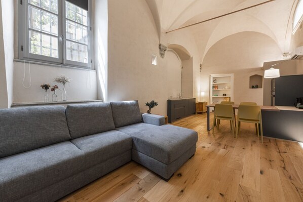 Un salon moderne avec un canapé d’angle gris, un plancher en bois, un coin repas avec des chaises jaunes et une cuisine dotée d’un comptoir foncé.