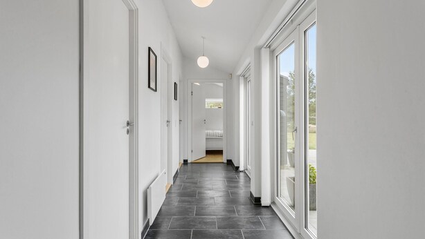 A modern hallway with white walls, dark tiled flooring, and large windows on one side. - 4 bedroom cozy home in ålbæk (Ålbæk)