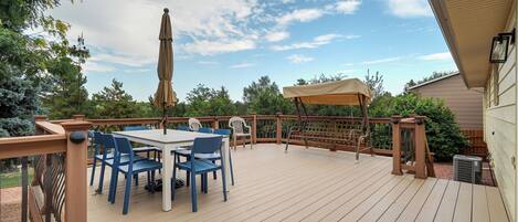 A wooden deck with a table and chairs, surrounded by greenery and a clear sky.