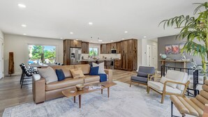 A spacious living area with a beige sofa, coffee table, and armchairs. A kitchen with wooden cabinets and modern appliances is visible in the background. - Colorado's Best (Colorado Springs)