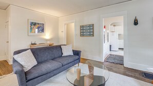 A modern living room with a grey sofa, a glass coffee table, and a white rug. There is a painting on the wall and a patterned artwork above the sofa.