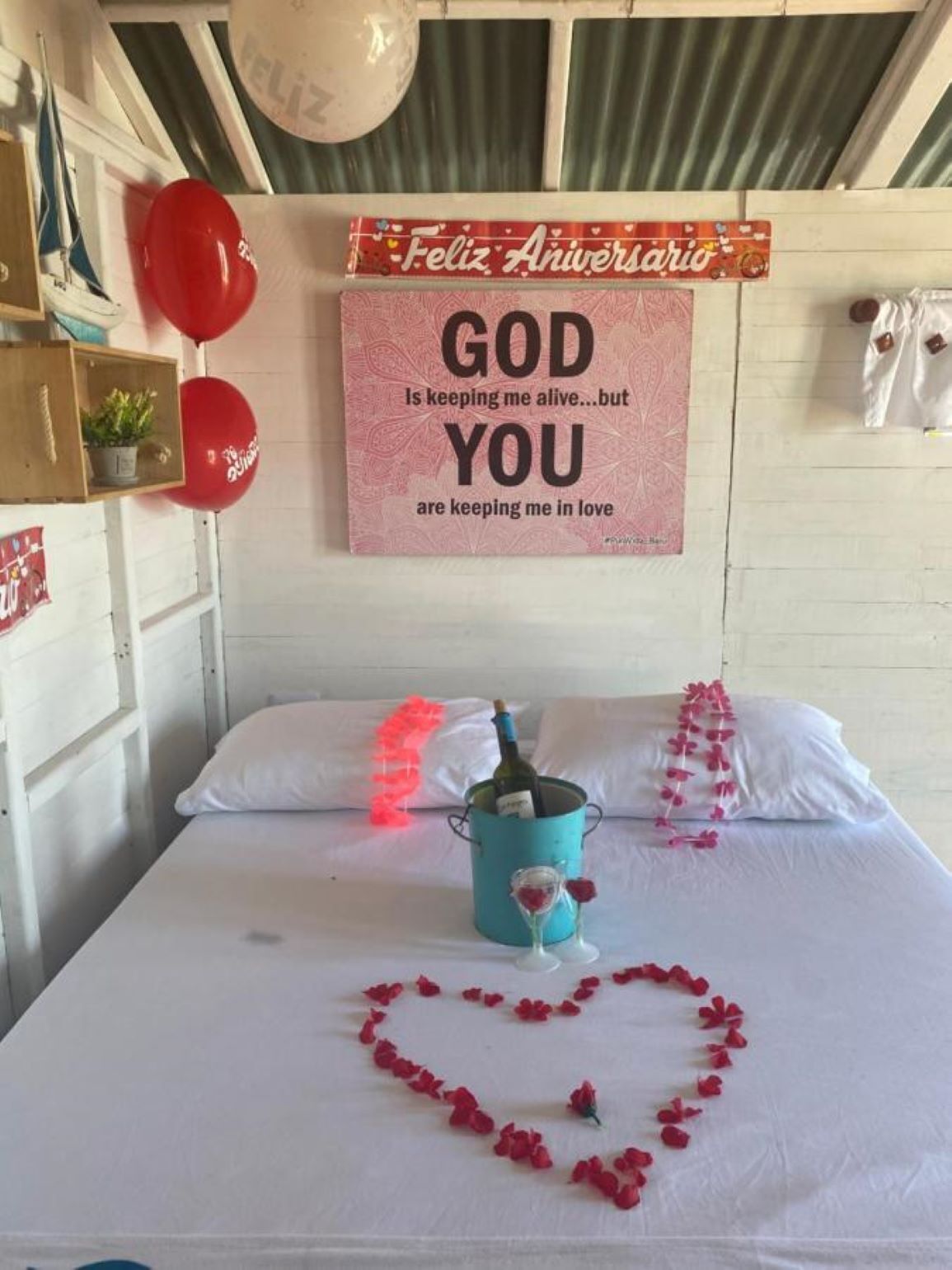 Un lit agrémenté d’un arrangement floral en forme de cœur, une bouteille de vin et une pancarte portant l’inscription « Feliz Aniversario » ainsi qu’un message sur l’amour et Dieu.