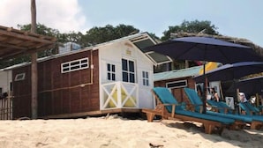 Een strandgebouw met rieten dak, een klein balkon en een rij blauwe ligstoelen onder een grote parasol.