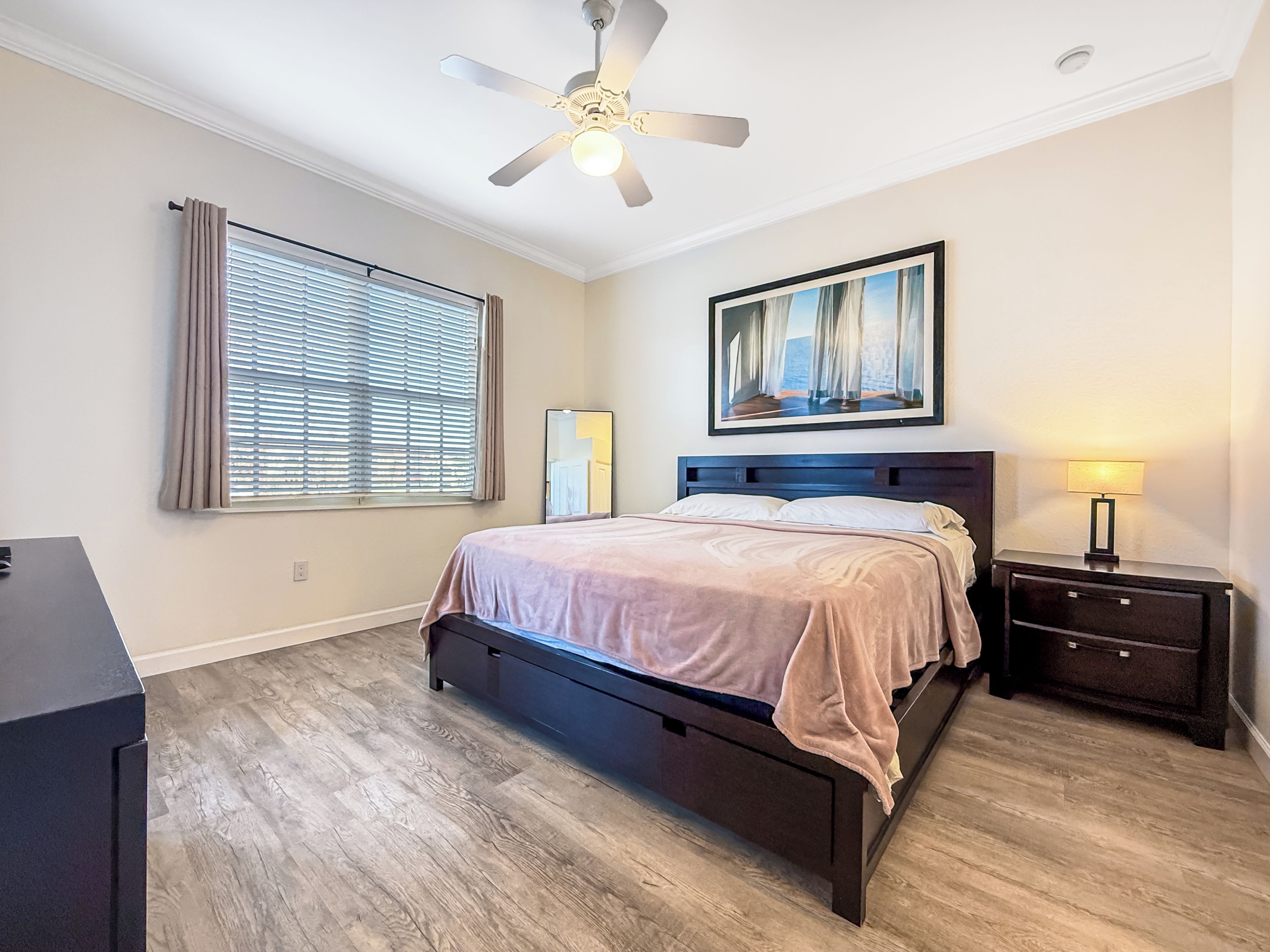 Une chambre à coucher comprenant un lit, une table de chevet, une lampe, un ventilateur de plafond, une fenêtre avec des stores et un tableau accroché au mur.