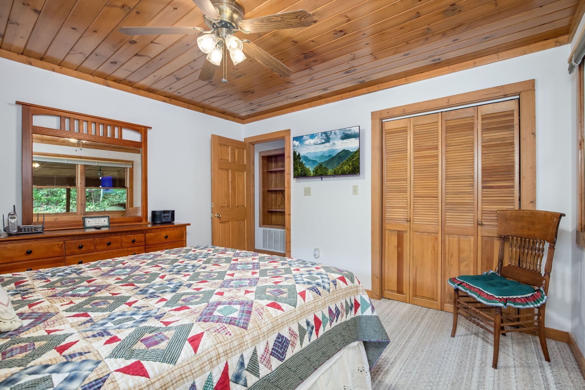 Un dormitorio con techo de madera, una cama con colcha de colores, cómoda de madera, una silla y una puerta que da a un baño.