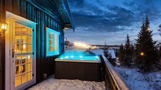 Una cabaña de madera con jacuzzi y vista a un paisaje nevado al anochecer.