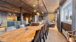 Un comedor amplio con una larga mesa de madera, varias sillas y un adorno central. Se observa una chimenea de piedra al fondo, y una ventana grande con cortinas en el lado derecho.