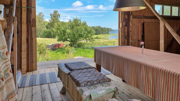 A wooden cabin with a large window overlooking a grassy area and a body of water. There is a bed with a patterned bedspread and a table with a floral arrangement. - Awesome home in Frändefors (Frändefors)
